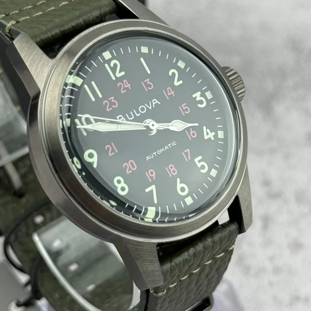 Bulova Hack 98A255 automatic mechanical watch with matte stainless steel case, black dial, red 24-hour numerals, and olive green genuine leather strap on wrist.
